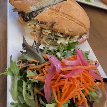 Commonwealth - Avocado Torta - A sandwich with toasted bread, filled with meat, vegetables, and sauce, is placed on a white rectangular platter. The sandwich appears to contain shredded lettuce, possibly meat, and other fillings. A side salad with mixed greens, shredded carrots, and pickled red onions is also on the platter. The setting suggests a casual dining environment, with additional dishes visible in the background. - American Near Me - Chicago, Illinois
