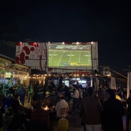 An outdoor bar area with a large screen showing a football game, surrounded by people sitting and standing, with some food trucks and string lights.