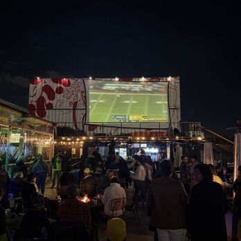 Metrobar DC - An outdoor bar area with a large screen showing a football game, surrounded by people sitting and standing, with some food trucks and string lights. - Bars Near Me - Washington, DC, District of Columbia