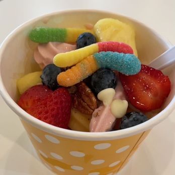 Yofruito - Mango and Strawberry yogurt with fruit and gummy's - A cup of frozen yogurt served in a yellow polka dot paper cup is topped with fresh strawberries, blueberries, gummy worms, and white chocolate chips. The yogurt appears to be flavored, with a spoon inserted into the cup, placed on a white table in a casual setting. - Ice Cream & Frozen Yogurt Near Me - Buffalo Grove, Illinois