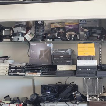 Technoland Computer Repairs - Where the magic happens - A shelf in a computer repair shop contains multiple electronic components and hardware items, including laptops, hard drives, and accessories. The top shelf holds various small electronic parts and tools, while the middle shelf features stacked laptops, some with labels and packaging. The bottom shelf has additional hardware, cables, and boxed items, indicating a collection of repair and replacement parts for computers and electronics. - IT Services & Computer Repair Near Me - Glendale, California