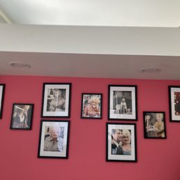 A pink interior wall features a collection of nine framed photographs of various people, arranged in a loose grid pattern. The frames are black with white mats, and the photos include portraits and candid shots. Reflections of window blinds are visible on some of the glass frames, indicating a nearby window. The wall is part of an indoor space, with a white ceiling and a small portion of a lamp visible in the lower right corner.