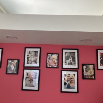 The Carrick - A pink interior wall features a collection of nine framed photographs of various people, arranged in a loose grid pattern. The frames are black with white mats, and the photos include portraits and candid shots. Reflections of window blinds are visible on some of the glass frames, indicating a nearby window. The wall is part of an indoor space, with a white ceiling and a small portion of a lamp visible in the lower right corner. - Hotels Near Me - Bisbee, Arizona