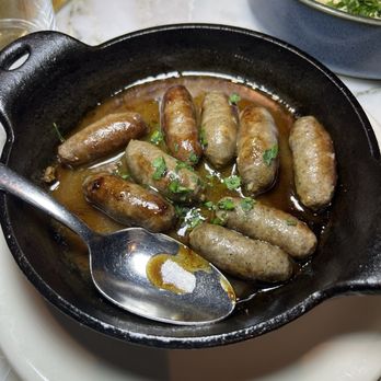 ilili DC - sausage - The image shows a black cast iron skillet filled with cooked sausages in a sauce, garnished with chopped green herbs. The skillet is placed on a white plate, and a spoon rests inside it. In the background, there is a bowl of a dish with rice and garnishes, and part of a glass is visible. The setting appears to be a restaurant table with a marble surface. - Lebanese Near Me - Washington, DC, District of Columbia