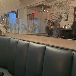 Christmas Decor - A mirror with snowflake ornaments and a silver ribbon decoration reflects a bar interior, showing patrons seated at the bar and a staff member preparing drinks. The scene includes Christmas decorations, framed pictures on the wall, and a cozy atmosphere with warm lighting.