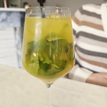 Tikka TapRoom - Mojito - A clear, tall wine glass holds a mojito cocktail with fresh mint leaves, lime slices, ice cubes, and a straw. The drink appears chilled and garnished with mint and lime, placed on a light-colored table. In the background, part of a person wearing a striped shirt is visible, along with a blurred indoor setting. - Indian Near Me - Lindenhurst, Illinois