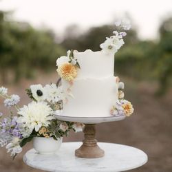 BTTRCRM - 2 tier vanilla cake on a stand with flowers in a vineyard.