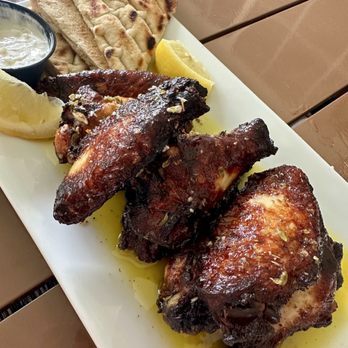 Pete's Taverna - Greek style wings - A rectangular white plate holds grilled chicken wings, flatbread, lemon wedges, and a small bowl of dipping sauce on a wooden table. - Cocktail Bars Near Me - Riverhead, New York