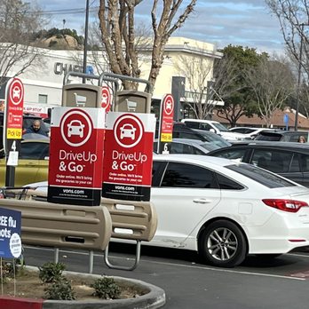 Vons - The image shows a parking lot outside a Vons grocery store in Riverside, CA. Two red and white signs indicate drive-up and go service, with a message about online ordering and grocery pickup. Several cars are parked, including a white sedan in the foreground. There are trees with bare branches and a building in the background. The scene depicts a typical grocery store parking area with designated pickup zones. - Grocery Near Me - Riverside, California