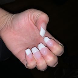 The image displays a hand with five fingers, each adorned with a French manicure. The nails have white tips and a clear or light pink base, and the hand is positioned against a dark background. The focus is on the well-groomed nails, highlighting the manicure's clean and polished appearance.