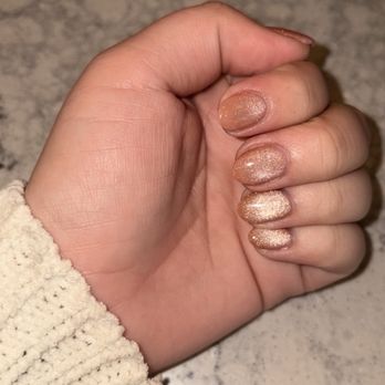 Nails By Nui - Full set, acrylic, magnetic polish - A photograph shows a person's left hand with neatly manicured nails painted in a shimmery, nude polish. The hand is positioned on a marble surface with a light, grayish pattern. The person is wearing a cream-colored, textured sweater sleeve. The image focuses on the nails and hand, with the background being the marble countertop. - Nail Technicians Near Me - Canton, Michigan