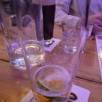 Ohio Brewing - The image shows a wooden table with multiple glasses of beverages, including a glass with a lime wedge and a clear liquid, a dark beer, and other empty or partially filled glasses. A person wearing a white shirt is partially visible in the background, and the setting appears to be a bar or restaurant with ambient lighting. - Breweries Near Me - Columbus, Ohio