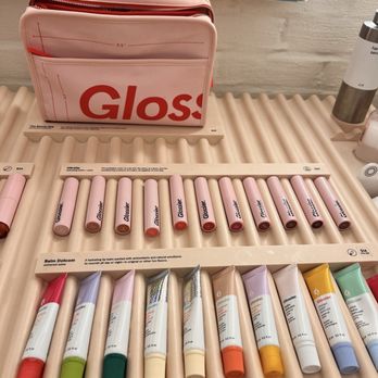 Glossier Chicago - The image shows a collection of Glossier lip balms and glosses arranged on a store counter, with a pink Glossier-branded bag behind them. - Cosmetics & Beauty Supply Near Me - Chicago, Illinois