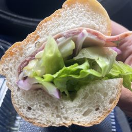 Italian Sub - A sandwich with lettuce, sliced onions, and deli meat is held in a person's hand inside a car, with a black interior visible.