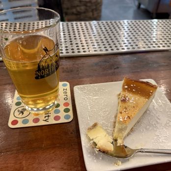 Fat Crow - My crème brûlée cheesecake - A glass of beer with a light golden color is positioned on a branded coaster on a wooden bar counter. Next to it, a slice of cheesecake with a caramelized top and powdered sugar is served on a white square plate, with a spoon resting on the plate. The background includes a metal bar mat and a blurred interior setting. - Pizza Near Me - Newfane, Vermont