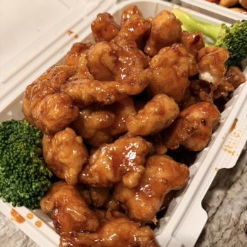 China Taste - Orange Chicken - A white takeout container holds pieces of orange chicken coated in a glossy, orange sauce, with some pieces showing a crispy exterior. Green broccoli florets are placed alongside the chicken as a garnish. The container is open, revealing the food inside, and is set on a marble countertop. Additional food containers are partially visible in the background. - Chinese Near Me - Windham, Maine