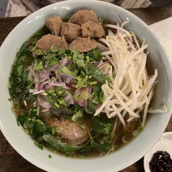 Pho Junkies - Custom pho: meatball, round eye, fatty brisket - A large bowl of pho contains meatballs, slices of beef, bean sprouts, chopped red onions, green onions, and fresh herbs such as cilantro. The broth is clear and the dish is presented on a wooden table. A small dish with black and red sauces is placed beside the bowl. - Food Trucks Near Me