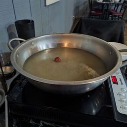 Chinese herb broth before adding anything else. - A large metal hot pot filled with Chinese herb broth is placed on an induction cooktop in a restaurant. The background shows dining tables and chairs.