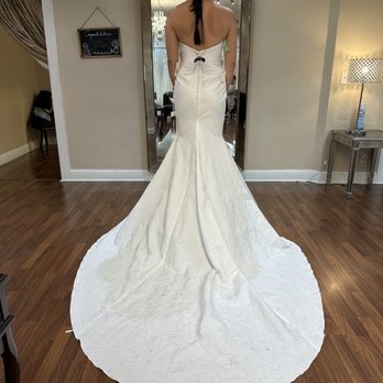Bo Lee - A woman wearing a strapless white wedding dress with a long, flowing train stands in front of a mirror, showing the back of her gown. Her dark hair is styled in a braid, and the setting appears to be a bridal boutique or fitting room with wooden flooring, a mirror, and decorative elements like a chalkboard sign and a lamp. - Bridal Near Me - Campbell, California