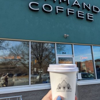 Command Coffee - A person with painted nails holds a disposable coffee cup with a mountain logo in front of Command Coffee, a modern coffee shop with large glass windows. The storefront features a teal exterior with prominent black and white signage. Reflections of trees and buildings are visible in the windows, and a neon 'Open' sign is illuminated inside. - Coffee & Tea Near Me - Indianapolis, Indiana