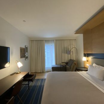 Canopy by Hilton Ithaca Downtown - The image shows a hotel room with a large bed positioned on the right side, with white bedding and pillows. To the left, a wall-mounted flat-screen TV is visible above a long desk or console. In the background, there is a sitting area with a chair, a small side table, and a floor lamp. The window is covered with cream-colored curtains, and the room has warm, ambient lighting. The decor includes framed artwork on the wall and a wooden accent wall behind the bed. - Hotels Near Me - Ithaca, New York