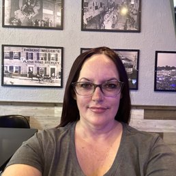 Stopped by for a pre-game soda before the game - A woman wearing glasses and a gray shirt is taking a selfie inside a restaurant or bar, with black-and-white framed photos on the wall behind her.
