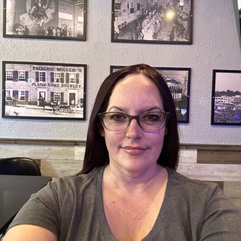 Kelly's Bleachers - Stopped by for a pre-game soda before the game - A woman wearing glasses and a gray shirt is taking a selfie inside a restaurant or bar, with black-and-white framed photos on the wall behind her. - Sports Bars Near Me - Milwaukee, Wisconsin