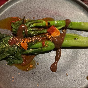 BearLeek - Gai Lan - The image shows a dish of grilled green vegetables, likely broccolini or asparagus, arranged on a gray plate. The vegetables are topped with a dark sauce, sprinkled with sesame seeds, and garnished with chili oil drops. The presentation emphasizes a combination of smoky, spicy, and savory flavors, typical of Asian-inspired cuisine. - American Near Me - Denver, Colorado