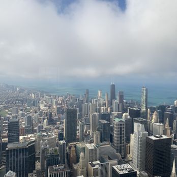 Skydeck Chicago - The image shows a high-angle view of Chicago's downtown area with numerous tall buildings and skyscrapers, including the Willis Tower. The cityscape extends toward Lake Michigan in the background, with a partly cloudy sky overhead. The photograph captures the urban density and architectural diversity of Chicago from an elevated perspective, likely from the Skydeck observation deck in the Willis Tower. - Observatories Near Me - Chicago, Illinois