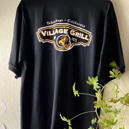 Village Grill bling. - A black shirt featuring a Village Grill logo from Tehachapi, California, is hanging on a wall. The logo includes a train illustration and the text "Village Grill" with the year 1991. The shirt is displayed against a textured wall, with a green leafy plant partially visible in the foreground.