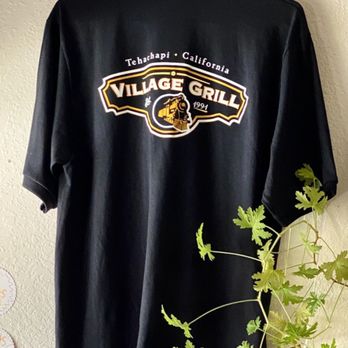 Village Grill - Village Grill bling. - A black shirt featuring a Village Grill logo from Tehachapi, California, is hanging on a wall. The logo includes a train illustration and the text "Village Grill" with the year 1991. The shirt is displayed against a textured wall, with a green leafy plant partially visible in the foreground. - Diners Near Me - Tehachapi, California