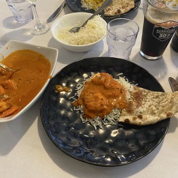 Mint - Indian Lakeside Dining - Bagari Jhinga and  Chicken Aam Wala - The image shows a table set with Indian food, including a black plate with rice topped with curry and a piece of naan bread. There is a white bowl of shredded cheese, a bowl of curry, and additional plates with rice and curry in the background. Drinks include a glass of white wine, a glass of water, and a glass of dark soda. The setting appears to be a casual dining environment. - Indian Near Me - Lee, Massachusetts