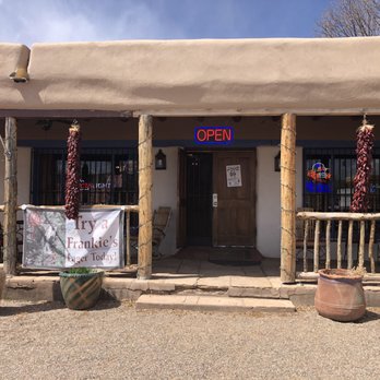 Frankie's at the Cassanova - The photo depicts the front exterior of Frankie's at the Cassanova, a restaurant with a Pueblo-style building featuring a beige stucco facade and wooden posts supporting a porch. An illuminated red 'OPEN' sign is displayed above the entrance, which has a wooden door. Decorative chili peppers hang on either side of the porch, and there are large ceramic pots with plants in front. An American flag is mounted on the left side of the porch, and a banner advertising a lager is also visible. The scene is set during daylight with a partly cloudy sky. - New American Near Me - Pecos, New Mexico
