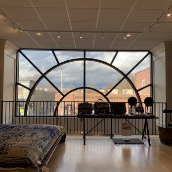 The Artisan's Bench - The view from the second floor window looking out over downtown Brighton! - The image shows the interior of a retail store with a large, decorative window featuring black geometric framing. The window allows natural light into the space and offers a view of buildings and a cloudy sky outside. Inside, there is a bed or rug on the left, and a table with various items, possibly merchandise, in front of the window. The ceiling has a grid pattern with track lighting. - Art Galleries Near Me - Brighton, Michigan