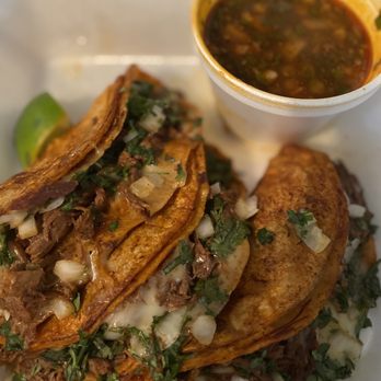El Compa - Yummy! - Two tacos with crispy corn tortillas filled with shredded meat, garnished with chopped onions and cilantro, served with a small cup of salsa, likely salsa verde or rojo, on a white tray. The tacos are presented with fresh ingredients and a side of spicy sauce. - Mexican Near Me - Birmingham, Alabama