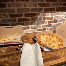 Pizza (Large) Garlic knots - A pizza box with cheese pizza, a garlic knots box, and a tray of garlic knots with dipping sauces are on a wooden table near a brick wall.