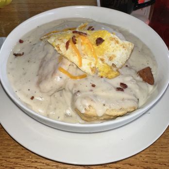 Market & Maple - A bowl of breakfast food contains eggs, gravy, and bacon pieces, with the eggs cooked sunny side up. The dish is served in a white bowl placed on a white plate, on a wooden table. A black mug and a red container are partially visible in the background. - Breakfast & Brunch Near Me - Dyer, Indiana