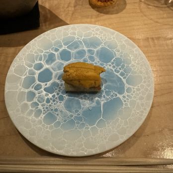 Hiden - Uni Toast with truffle salt - A piece of uni (sea urchin) sushi is centered on a round plate featuring a blue and white bubble pattern. The plate is on a wooden table, with a glass and a metallic bowl partially visible in the background. The setting suggests a dining experience, likely at a sushi or omakase restaurant. - Japanese Near Me - Miami, Florida