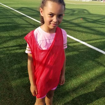 Q2 Stadium - Thing Two in the practice Jersey - A young girl with curly hair stands on a well-maintained grass soccer field, wearing a pink shirt, pink shorts, black sneakers, and a red training vest. The field has white boundary lines, and there are soccer goals, parked cars, and a building in the background. Shadows from nearby objects are cast on the grass, indicating sunny weather. - Stadiums & Arenas Near Me - Austin, Texas