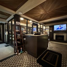 Retail counter and humidor. - The image shows the interior of a cigar shop or lounge with a reception or checkout counter in the center. Behind the counter are glass display cases filled with cigar products. A wall-mounted TV is visible on the right side, displaying a sports or news program. The space has a patterned tile floor, dark wood accents, and a wooden ceiling with geometric paneling. There is a ladder-style shelf next to the counter with cleaning supplies and a door on the left side leading to another area.