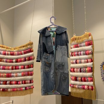 MOWA - St Kate - The image shows two textile art pieces with colorful, patterned fabric rolls, each bordered with yellow fringe, hanging from chains. Between them, a denim jacket with multiple pockets and a hood is displayed on a purple hanger. The background is a plain, light-colored wall with a small decorative piece on the right side. The display appears to be part of an art or fashion exhibit. - Art Galleries Near Me - Milwaukee, Wisconsin