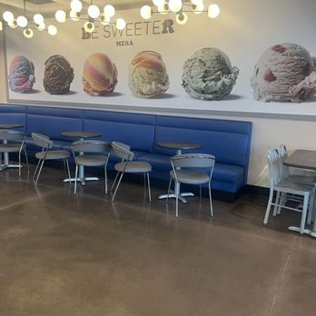 Baskin-Robbins - seating area - The image shows an indoor seating area at Baskin-Robbins in Mesa, with a long blue upholstered booth along the wall and several metal chairs with gray seats arranged around small round tables. The wall behind the seating has a large mural of ice cream scoops and the text 'BE SWEETER' and 'B R' in colorful letters. The ceiling has modern lighting fixtures with multiple round bulbs, and the floor is polished concrete. - Ice Cream & Frozen Yogurt Near Me - Mesa, Arizona