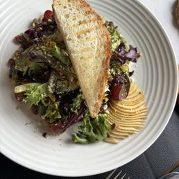Field Green & Pear Salad - A salad featuring mixed greens, grape tomatoes, shredded cheese, and chopped nuts is presented on a white plate. A large slice of toasted bread rests on top of the salad. The dish is placed on a dark surface with a fork and knife nearby, and part of a paper napkin is visible in the background.