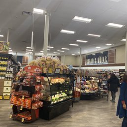 people shopping in a grocery store - The image shows the interior of a grocery store with a focus on the bread and rolls section. Several shoppers are browsing the aisles, which are stocked with various baked goods, packaged bread, and other grocery items. The store has bright lighting, a clean and organized layout, and visible signage indicating different sections. The flooring appears to be wood or laminate, and there are shelves and displays arranged for customer convenience.