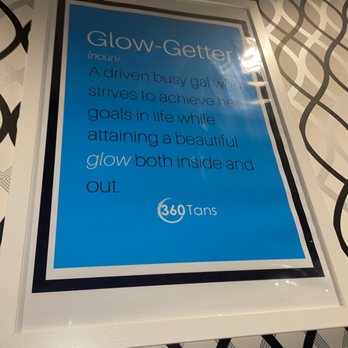 360 Tans - A framed sign with a blue background and white text provides a definition and description of Glow-Getter, highlighting qualities of a driven individual striving for goals and inner and outer glow. The sign includes the 360 Tans logo at the bottom and is mounted on a wall with a black and white patterned background. - Spray Tanning Near Me - Sunset Valley, Texas