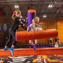 Altitude Trampoline Park - Appleton - A boy and a girl stand on a narrow, padded beam facing each other while holding large, colorful foam jousting sticks...
