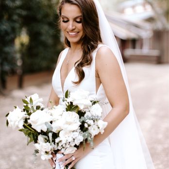 Events By B+D - Bridal Bouquet - A smiling bride in a white wedding gown with a veil holds a large bouquet of white flowers outdoors, with blurred trees and sunlight in the background. - Party & Event Planning Near Me