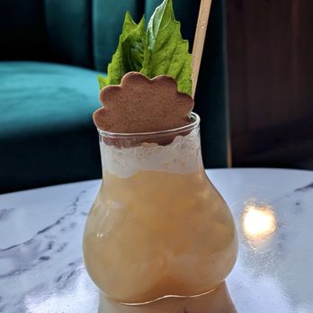 Public Parking - A cocktail with foam and a light color, garnished with fresh green herbs and a cookie, placed on a marble table in a cozy indoor setting. - Bars Near Me - Madison, Wisconsin