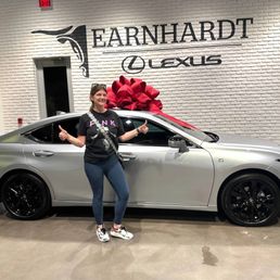 A woman stands in front of a silver Lexus car with a large red bow on top, inside the Earnhardt Lexus dealership in Phoenix. She is smiling and giving a thumbs-up, wearing a black T-shirt, jeans, and sneakers. The background features a white brick wall with the Earnhardt Lexus logo and signage, along with some equipment and a doorway. The scene suggests a vehicle delivery or celebration event.