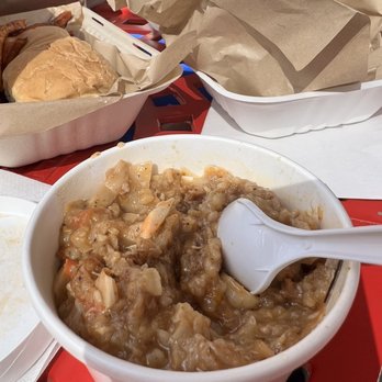 Herban Vegans - Gumbo - A bowl of vegan gumbo with a white spoon is on a red table, with paper-wrapped food items in the background. - Vegan Near Me - Cincinnati, Ohio